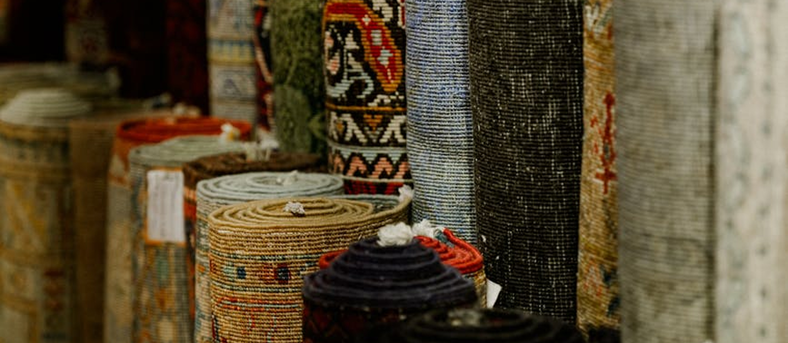 Rows of handwoven carpets and textiles rolled and standing — representing the artisan women at the heart of Rita Industries